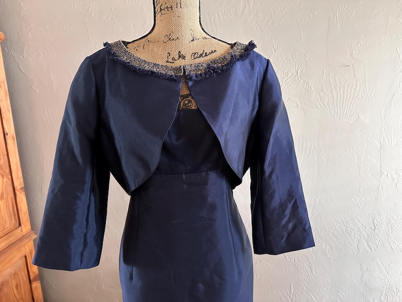 May include: A navy blue dress with a matching bolero jacket. The dress has a fitted silhouette, and the jacket features three-quarter sleeves and a decorative trim around the neckline. The outfit is displayed on a mannequin.