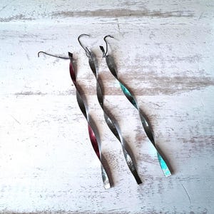 Puede incluir: Tres piezas decorativas de metal retorcido con un gancho en la parte superior. El metal tiene un acabado plateado con toques de rojo y turquesa. Las piezas están sobre una superficie blanca texturizada.