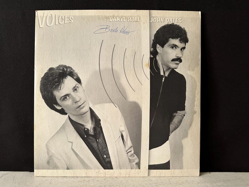 May include: A black and white album cover with the title "Voices" and the names "Daryl Hall" and "John Oates". The album cover features two men, one with short hair and a suit jacket, and the other with longer hair and a dark jacket. The album cover has a sound wave design.