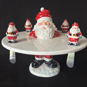 Può includere: Un porta torta di Natale in ceramica bianca con una statuetta di Babbo Natale in cima. Il supporto ha quattro piccole statuine di Babbo Natale intorno al bordo e le parole "Time for a little something" sulla base.