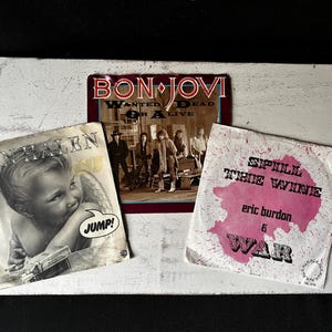 Puede incluir: Tres discos de vinilo vintage, uno rosa, uno blanco y uno rojo, con los nombres de las bandas "Van Halen", "Bon Jovi" y "Eric Burdon & War".