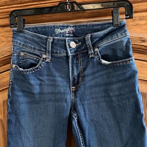 Pode incluir: Cal&ccedil;as de ganga azuis Wrangler, corte bota, penduradas num cabide preto. As cal&ccedil;as de ganga t&ecirc;m fecho de bot&atilde;o, bolsos frontais e traseiros e costuras decorativas. O log&oacute;tipo Wrangler &eacute; vis&iacute;vel na cintura.