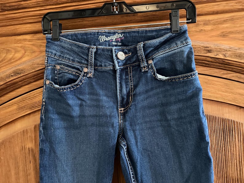 Pode incluir: Cal&ccedil;as de ganga azuis Wrangler, corte bota, penduradas num cabide preto. As cal&ccedil;as de ganga t&ecirc;m fecho de bot&atilde;o, bolsos frontais e traseiros e costuras decorativas. O log&oacute;tipo Wrangler &eacute; vis&iacute;vel na cintura.