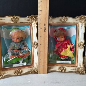 May include: Three framed pictures of dolls dressed as fairy tale characters. The dolls are dressed as Little Bo Peep, Red Riding Hood, and a third character. The frames are gold and white.