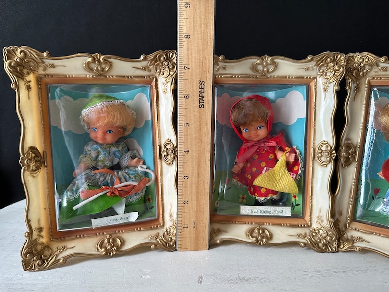 May include: Three framed pictures of dolls dressed as fairy tale characters. The dolls are dressed as Little Bo Peep, Red Riding Hood, and a third character. The frames are gold and white.