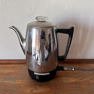 May include: A vintage General Electric electric percolator coffee pot with a polished chrome finish, a black plastic handle, and a clear glass lid knob. The base is black with a control dial labeled MILD and STRONG, and a black power cord attached.