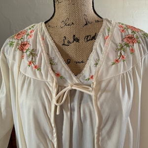 Könnte beinhalten: Ein weißes, langärmliges Gewand mit V-Ausschnitt und Blumenstickerei. Das Gewand hat einen Bindegürtel in der Taille und einen Spitzenbesatz am Ausschnitt.