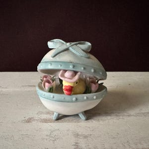 May include: A ceramic Easter egg-shaped trinket box in shades of white and light blue, with a duckling wearing a pink hat inside. Pink roses and a blue bow decorate the egg. The duckling has a yellow body and a red beak.