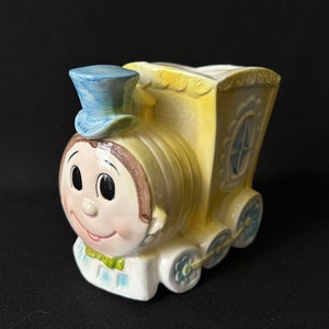 Può includere: Un vaso da fiori in ceramica giallo a forma di treno con un cappello blu e un viso sorridente. Il treno ha un piccolo finestrino e ruote con fiori blu.