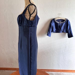 May include: A navy blue dress with a matching bolero jacket. The dress is displayed on a mannequin and has a fitted bodice and a straight skirt. The bolero jacket is short-sleeved and has a beaded neckline. The dress and jacket are made of a shiny fabric.
