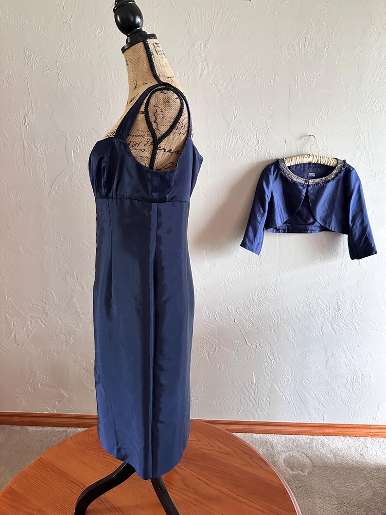 May include: A navy blue dress with a matching bolero jacket. The dress is displayed on a mannequin and has a fitted bodice and a straight skirt. The bolero jacket is short-sleeved and has a beaded neckline. The dress and jacket are made of a shiny fabric.