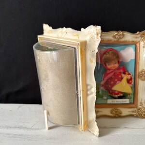 May include: A vintage white picture frame with a gold trim and a glass front. The frame is holding a picture of a red-haired doll wearing a red dress and a yellow basket. The text "Red Riding Hood" is visible in the picture.
