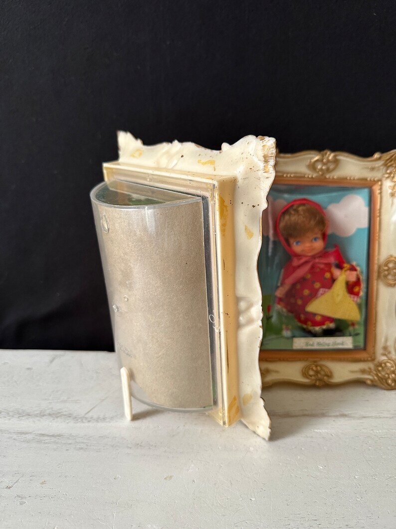 May include: A vintage white picture frame with a gold trim and a glass front. The frame is holding a picture of a red-haired doll wearing a red dress and a yellow basket. The text "Red Riding Hood" is visible in the picture.
