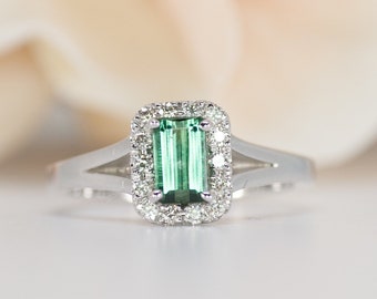 Puede incluir: Un anillo de plata con una piedra preciosa verde de talla esmeralda rodeada por un halo de diamantes más pequeños.