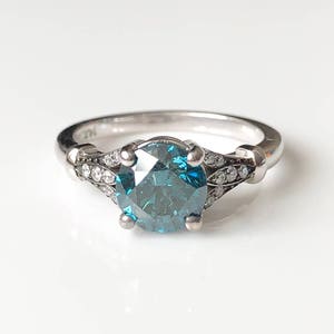 K&ouml;nnte beinhalten: Ein silberner Ring mit einem gro&szlig;en, runden, teal-farbenen Edelstein als Mittelpunkt. Das Band ist mit kleineren, klaren Edelsteinen verziert. Der Ring ist vor einem wei&szlig;en Hintergrund platziert, der seine Details hervorhebt.