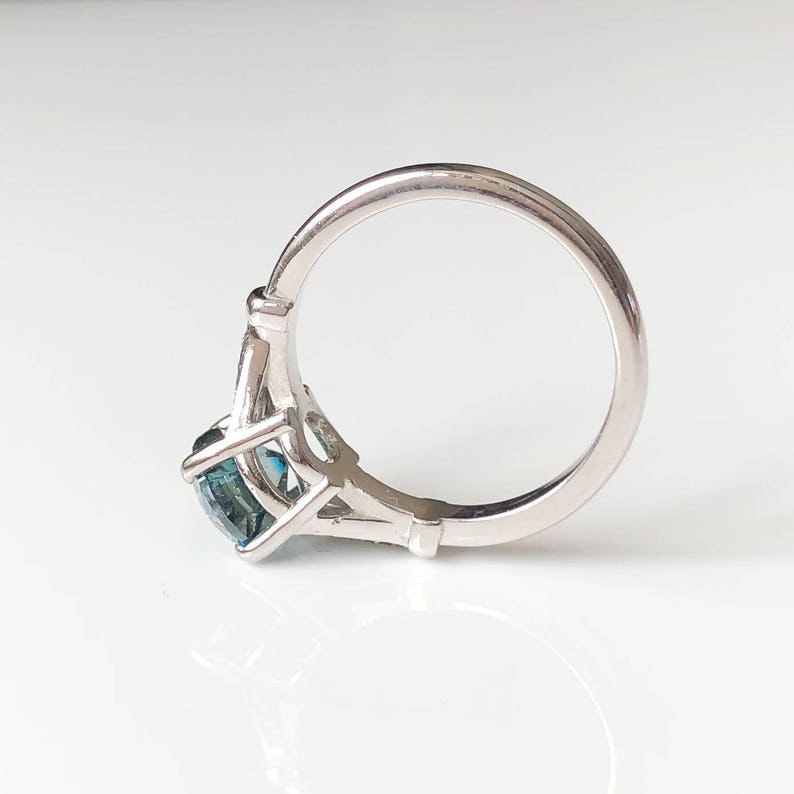 K&ouml;nnte beinhalten: Ein silberner Ring mit einem gro&szlig;en, quadratisch geschliffenen, teal-farbenen Edelstein. Der Ring hat ein glattes, poliertes Band, und der Edelstein wird von filigranen Zinken gehalten. Der Ring ist vor wei&szlig;em Hintergrund.