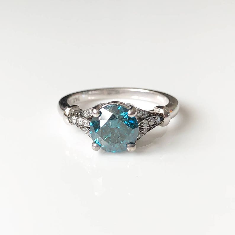 K&ouml;nnte beinhalten: Ein silberner Ring mit einem gro&szlig;en, runden, teal-farbenen Edelstein als Mittelpunkt. Das Band ist mit kleineren, klaren Edelsteinen verziert. Der Ring ist vor einem wei&szlig;en Hintergrund platziert, der seine Details hervorhebt.