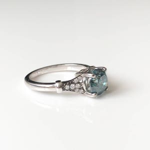 K&ouml;nnte beinhalten: Ein silberner Ring mit einem gro&szlig;en, runden, teal-farbenen Edelstein als Mittelpunkt. Das Band ist mit kleinen, klaren Akzentsteinen und dekorativen Elementen versehen. Der Ring ist vor einem wei&szlig;en Hintergrund platziert.
