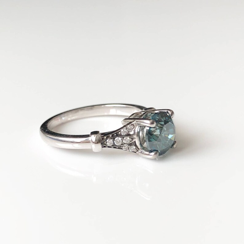 K&ouml;nnte beinhalten: Ein silberner Ring mit einem gro&szlig;en, runden, teal-farbenen Edelstein als Mittelpunkt. Das Band ist mit kleinen, klaren Akzentsteinen und dekorativen Elementen versehen. Der Ring ist vor einem wei&szlig;en Hintergrund platziert.