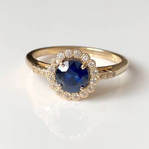 K&ouml;nnte beinhalten: Ein goldener Ring mit einem blauen Saphir in der Mitte, umgeben von einem Heiligenschein aus kleineren Diamanten.