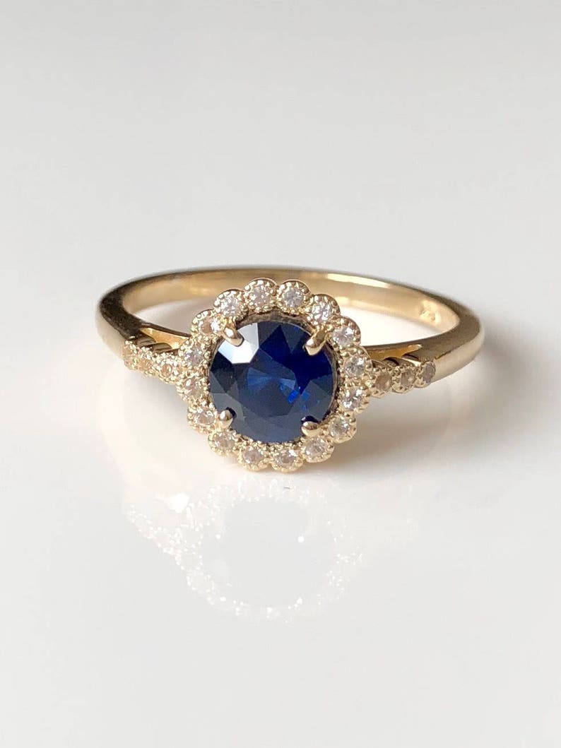 K&ouml;nnte beinhalten: Ein goldener Ring mit einem blauen Saphir in der Mitte, umgeben von einem Heiligenschein aus kleineren Diamanten.