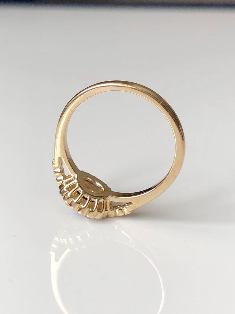 K&ouml;nnte beinhalten: Ein goldener Ring mit einem einzigen, runden, klaren Edelstein, der in der Mitte sitzt. Die Ringschiene ist mit einer Reihe kleiner, runder Edelsteine auf jeder Seite des Mittelst&uuml;cks verziert.