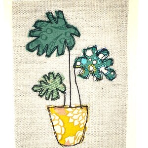May include: A hand-stitched fabric art piece depicting a potted plant with green leaves and a yellow patterned pot on a gray background.