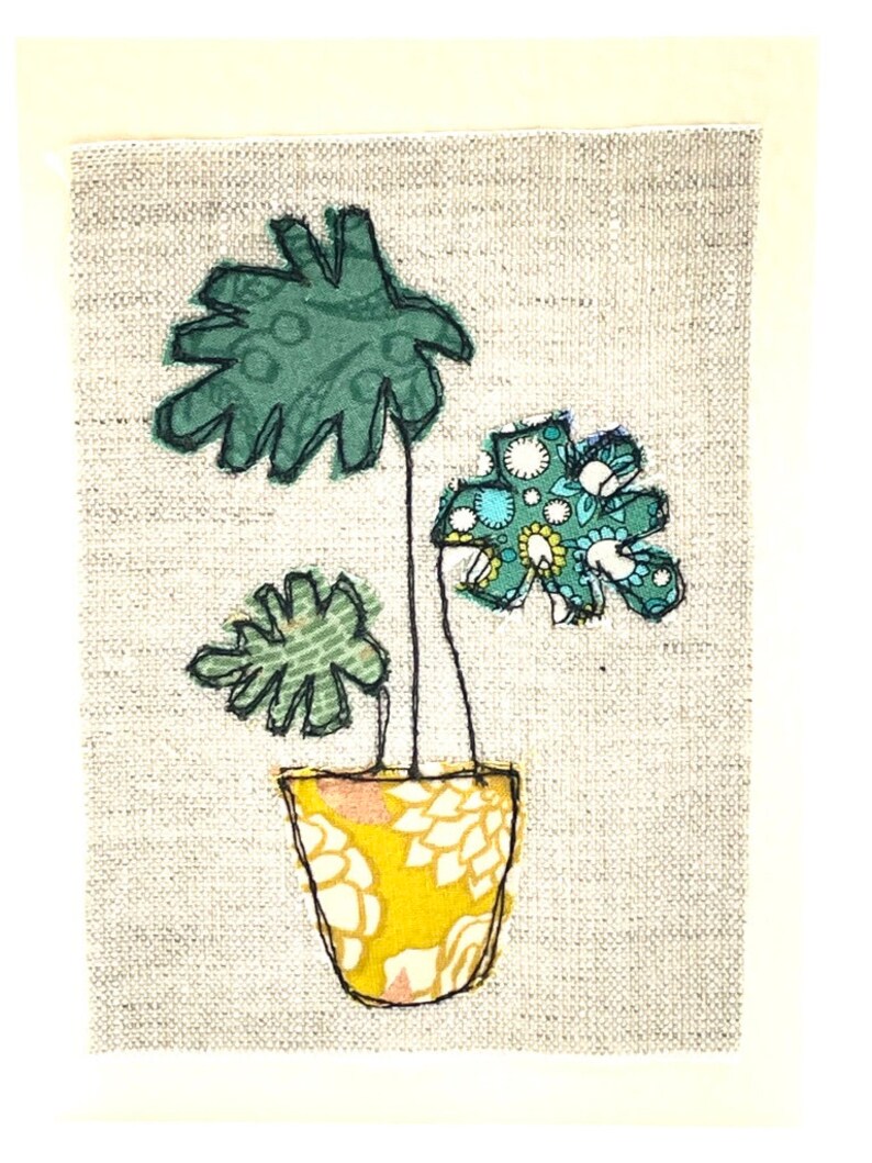 May include: A hand-stitched fabric art piece depicting a potted plant with green leaves and a yellow patterned pot on a gray background.