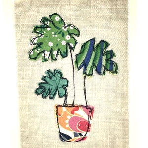 May include: A green and blue fabric plant with white flowers in a colorful patterned pot on a beige background.