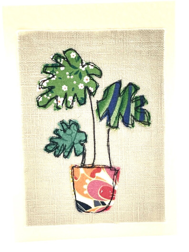 May include: A green and blue fabric plant with white flowers in a colorful patterned pot on a beige background.