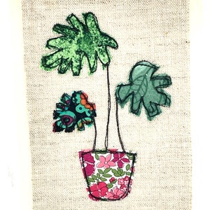 May include: A green fabric plant with three leaves in a pink and floral patterned fabric pot on a beige linen background.