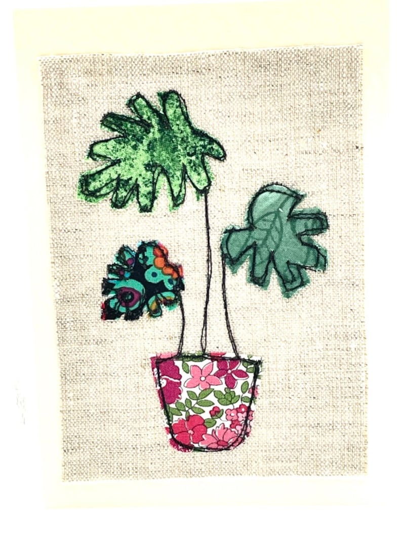 May include: A green fabric plant with three leaves in a pink and floral patterned fabric pot on a beige linen background.