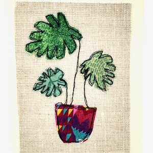 May include: A green plant with three leaves in a colorful patterned pot. The plant is stitched onto a beige fabric background.