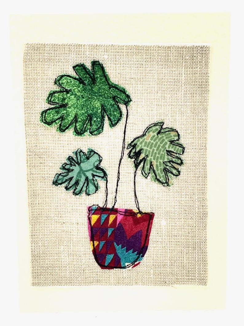 May include: A green plant with three leaves in a colorful patterned pot. The plant is stitched onto a beige fabric background.