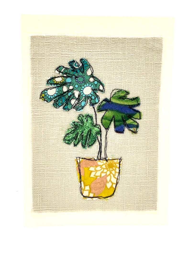 May include: A hand-stitched embroidery of a potted plant with green, blue, and white leaves on a beige background.