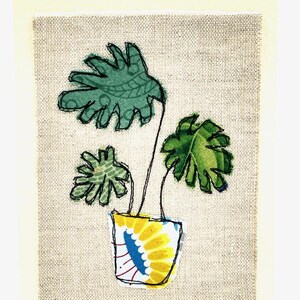 May include: A hand-stitched artwork featuring a green plant with large leaves in a yellow pot with a blue and white pattern on a beige linen background.