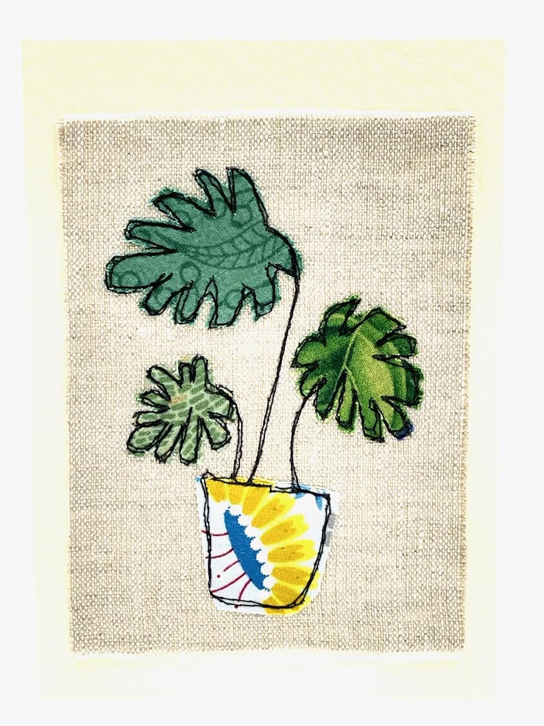 May include: A hand-stitched artwork featuring a green plant with large leaves in a yellow pot with a blue and white pattern on a beige linen background.