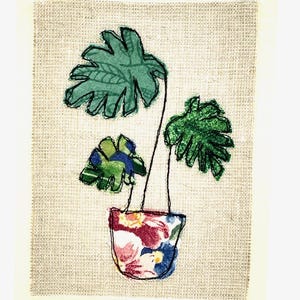 May include: A green fabric plant with three leaves in a floral patterned fabric pot on a beige background.