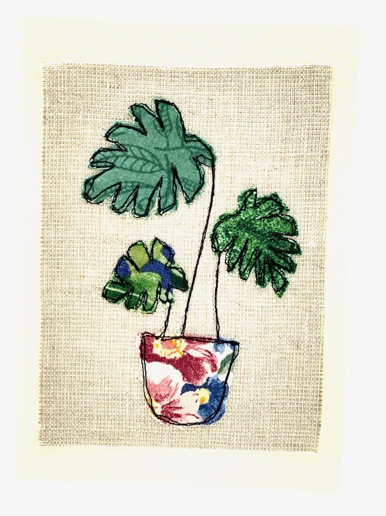 May include: A green fabric plant with three leaves in a floral patterned fabric pot on a beige background.