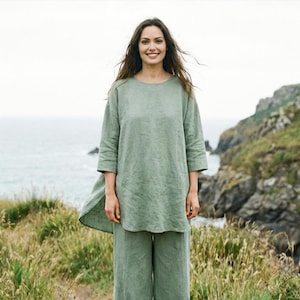 Può includere: Una donna indossa un completo in lino verde salvia, composto da una tunica con maniche a tre quarti e pantaloni ampi. La tunica ha uno scollo rotondo e una vestibilità ampia. Lo sfondo mostra un paesaggio costiero con mare e scogliere.