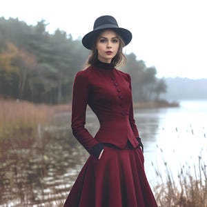 May include: A woman in a burgundy outfit, comprising a skirt and jacket, with black gloves and a black hat. The jacket has a high collar and button details. The background features a lake and trees.