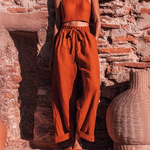 May include: A rust-coloured linen two-piece set, comprising a crop top and high-waisted trousers with a drawstring waist. The trousers feature pockets and are cuffed at the ankles. The outfit is displayed against a textured stone wall.