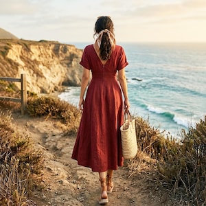 May include: A woman in a rust-colored midi dress walks along a coastal path, carrying a woven tote bag. The dress features a V-neck and short sleeves. The background shows a cliffside, ocean, and a wooden fence.