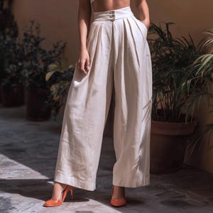 May include: Cream-colored wide-leg pants with a high waist and button closure. The pants have pleats and pockets. The model is wearing orange heels. The background includes potted plants and a neutral-toned wall.