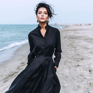May include: A woman in a long black dress with a button-down top and a tie-waist skirt. The dress has long sleeves and a mandarin collar. She is wearing gold earrings and standing on a sandy beach with the sea in the background.