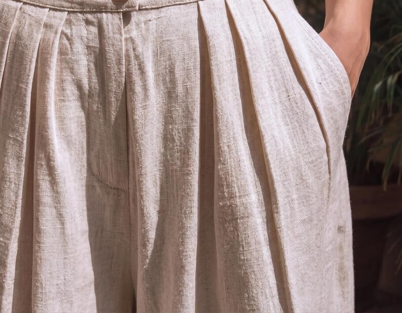 May include: Close-up of beige linen pants with pleats and a pocket. The fabric has a textured appearance. The pants are high-waisted and appear to be made of a lightweight material, suitable for warm weather.