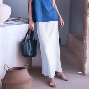 Puede incluir: Una mujer con una blusa azul y una falda blanca, sosteniendo un bolso tote negro. El atuendo se combina con sandalias. Una cesta tejida y un jarrón están en el fondo.