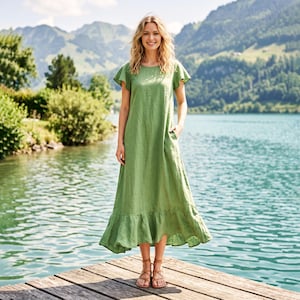 May include: A woman in a long, olive green linen dress with flutter sleeves and a ruffled hem, standing on a wooden dock. The dress has pockets and is worn with brown sandals. The background shows a lake and mountains.