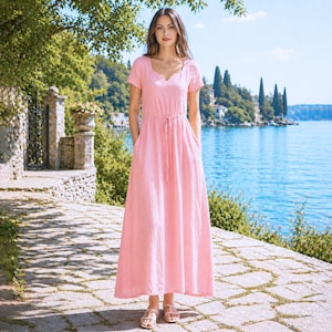 May include: A light pink maxi dress with short sleeves and a V-neckline. The dress features a drawstring waist and pockets. The model is wearing sandals and standing on a stone path near a lake.