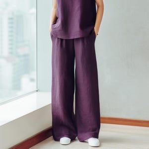 May include: A two-piece outfit comprising wide-leg trousers and a sleeveless top in a deep purple shade. The trousers have a loose fit, and the top has a simple, flowing design. The ensemble is completed with white trainers, creating a relaxed, comfortable appearance.